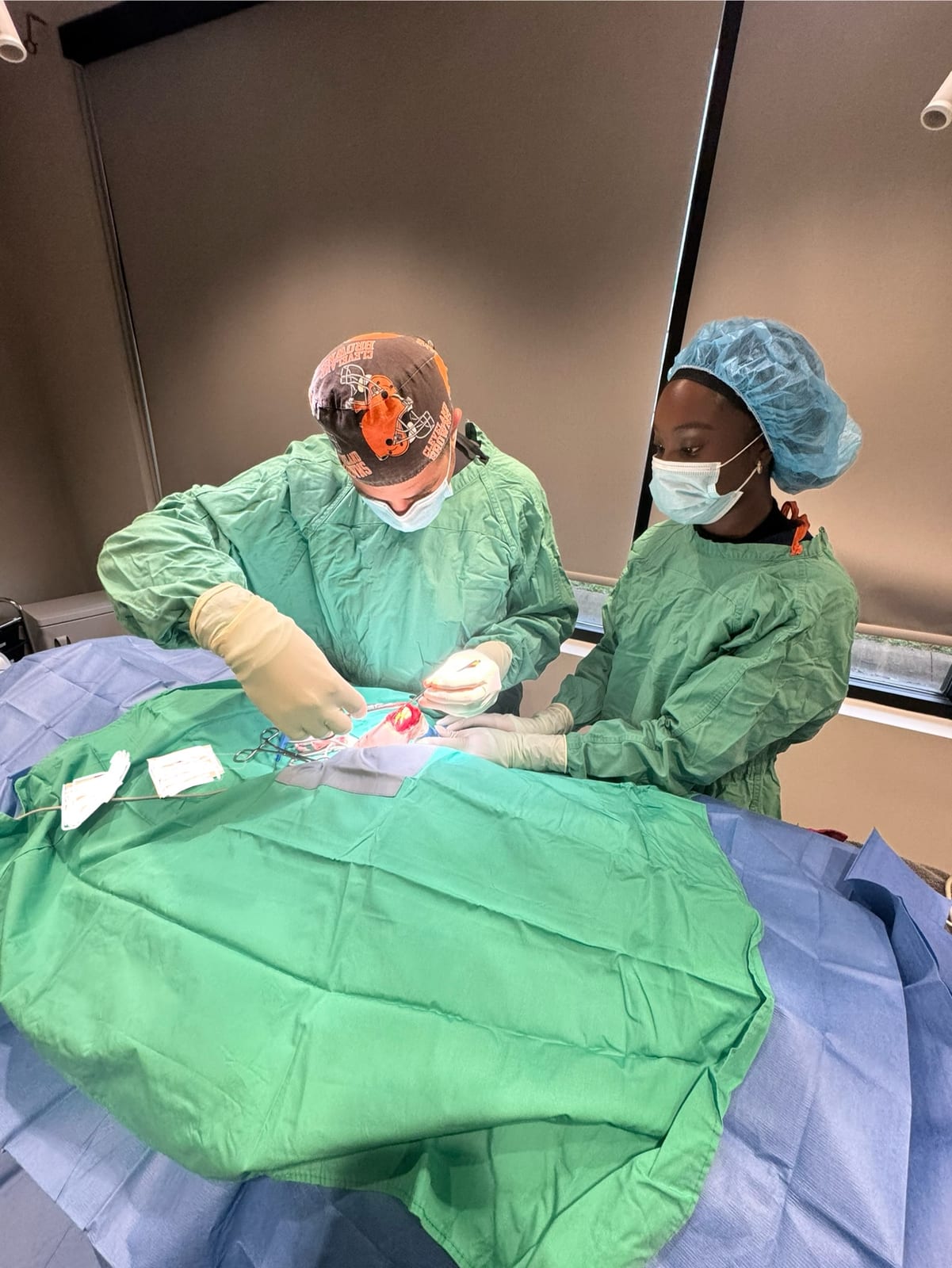 Two people in green surgical gowns, caps and masks working over a green and blue draped table