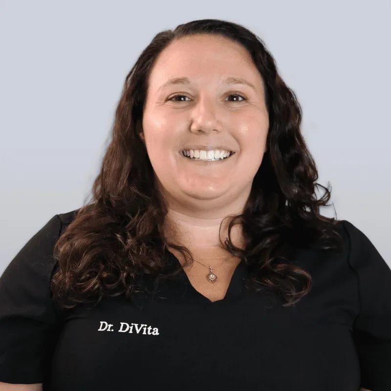 Smiling woman with long curly brown hair in a black scrub top with embroidered white text on a pale grey background