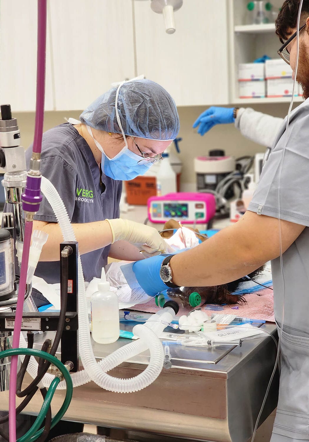 Two people in scrub caps and masks working at a steel table with tubing and a monitor