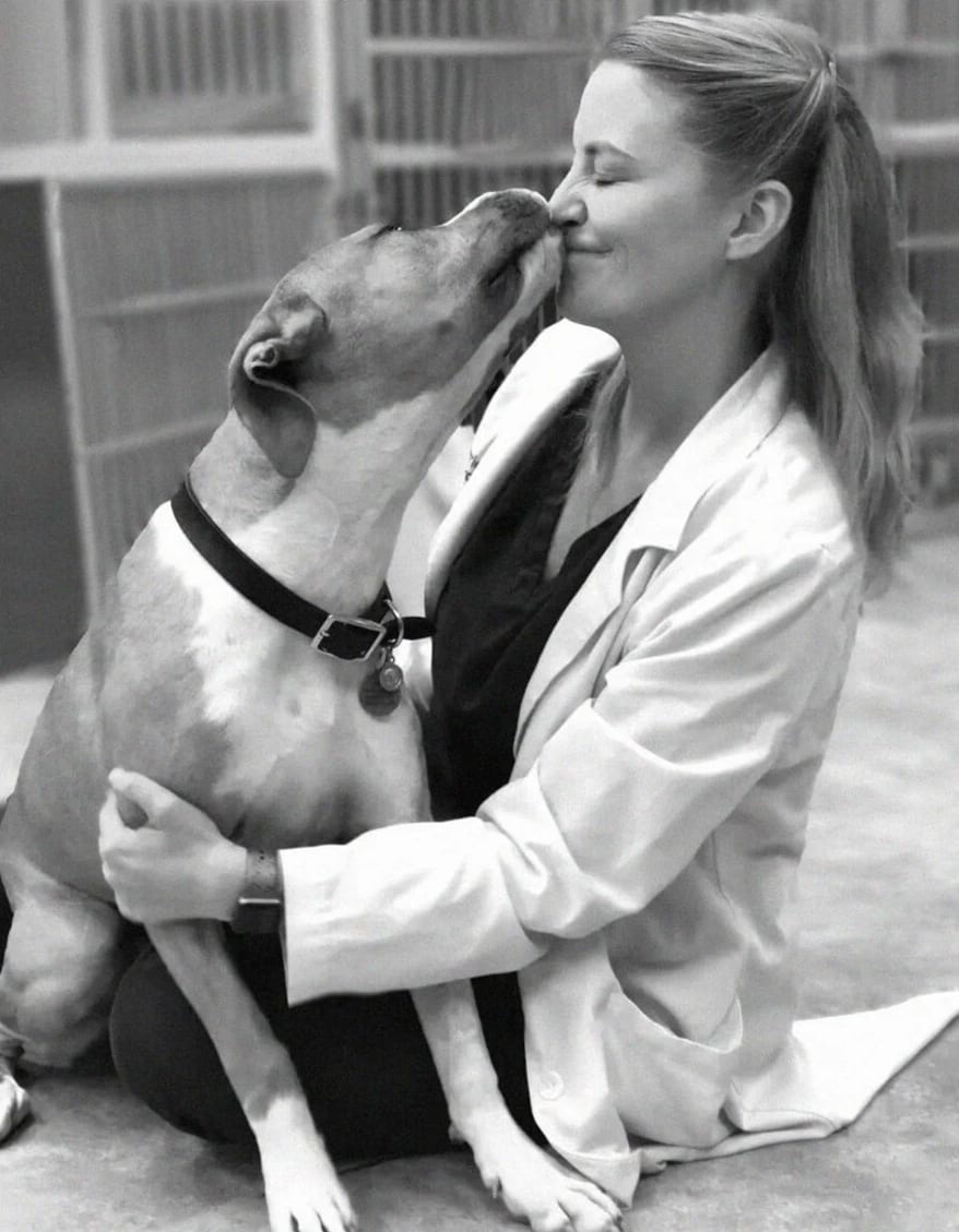 Black and white photo of a person in a white coat sitting on the floor as a dog licks their face