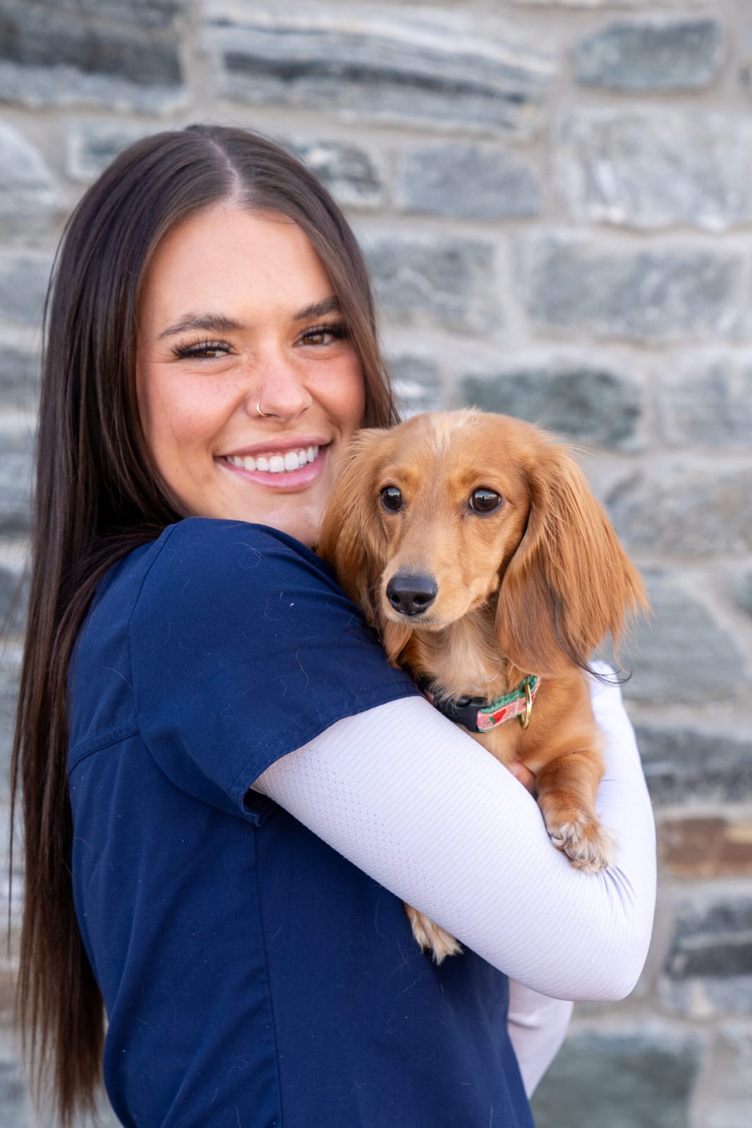 Smiling person in a navy scrub top holding a small tan dog in front of a stone wall