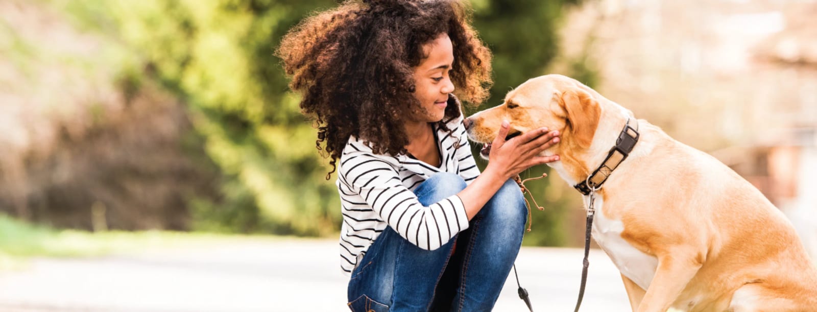 Young person in a striped top crouching to pet a tan dog on a sunny path