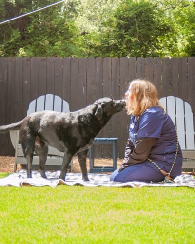 Person in navy scrubs sitting on a blanket in a yard as a black dog licks their face