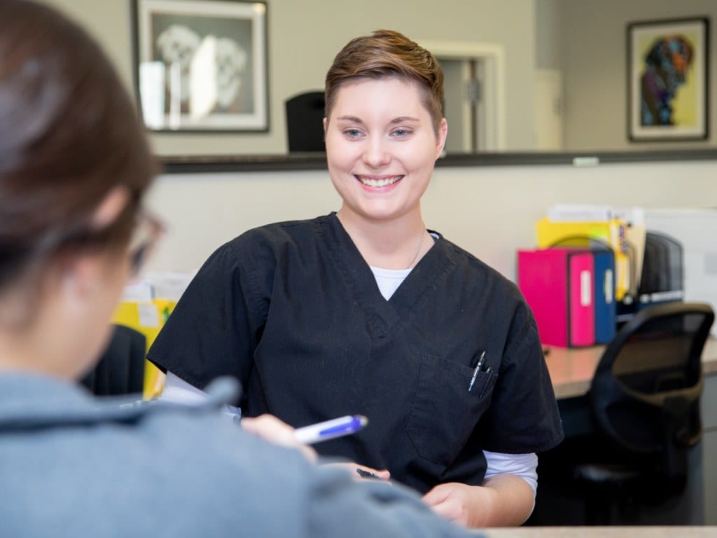 Smiling person in black scrubs at a reception counter talking to a person in the foreground