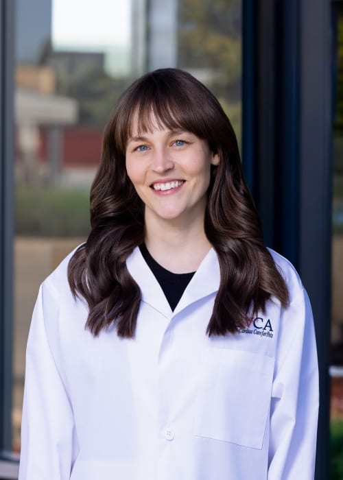 Smiling woman with long wavy brown hair wearing a white coat, standing in front of glass windows