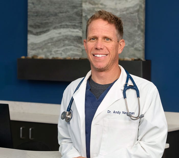 Smiling man with short hair in a white coat with a blue stethoscope in front of a blue wall and stone panel