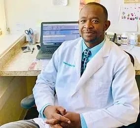 Smiling person in a white coat and patterned tie sitting at a desk with a laptop