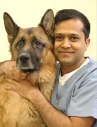 Smiling man in grey-blue scrubs with his arms around a large tan and black dog