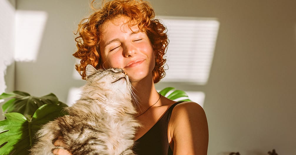 Woman with curly red hair and closed eyes being nuzzled by a fluffy cat beside a leafy plant
