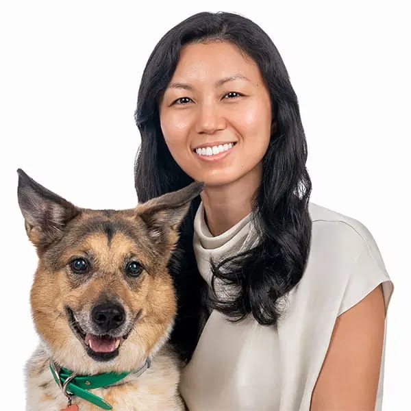 Smiling woman with long dark hair beside a tan and black dog in a green collar on a white background