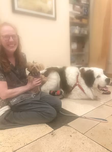 Smiling woman sitting on a tiled floor holding a small dog beside a large black and white dog