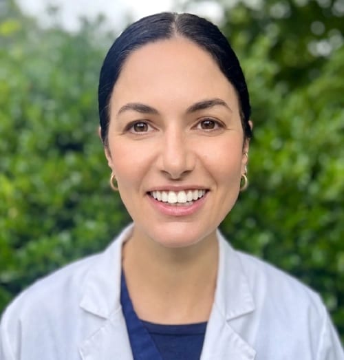 Portrait of a smiling woman with dark hair pulled back, wearing a white coat over navy scrubs