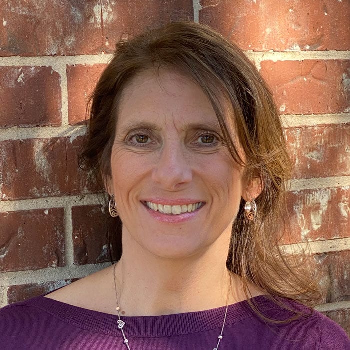 Smiling woman with long brown hair in a purple top in front of a red brick wall