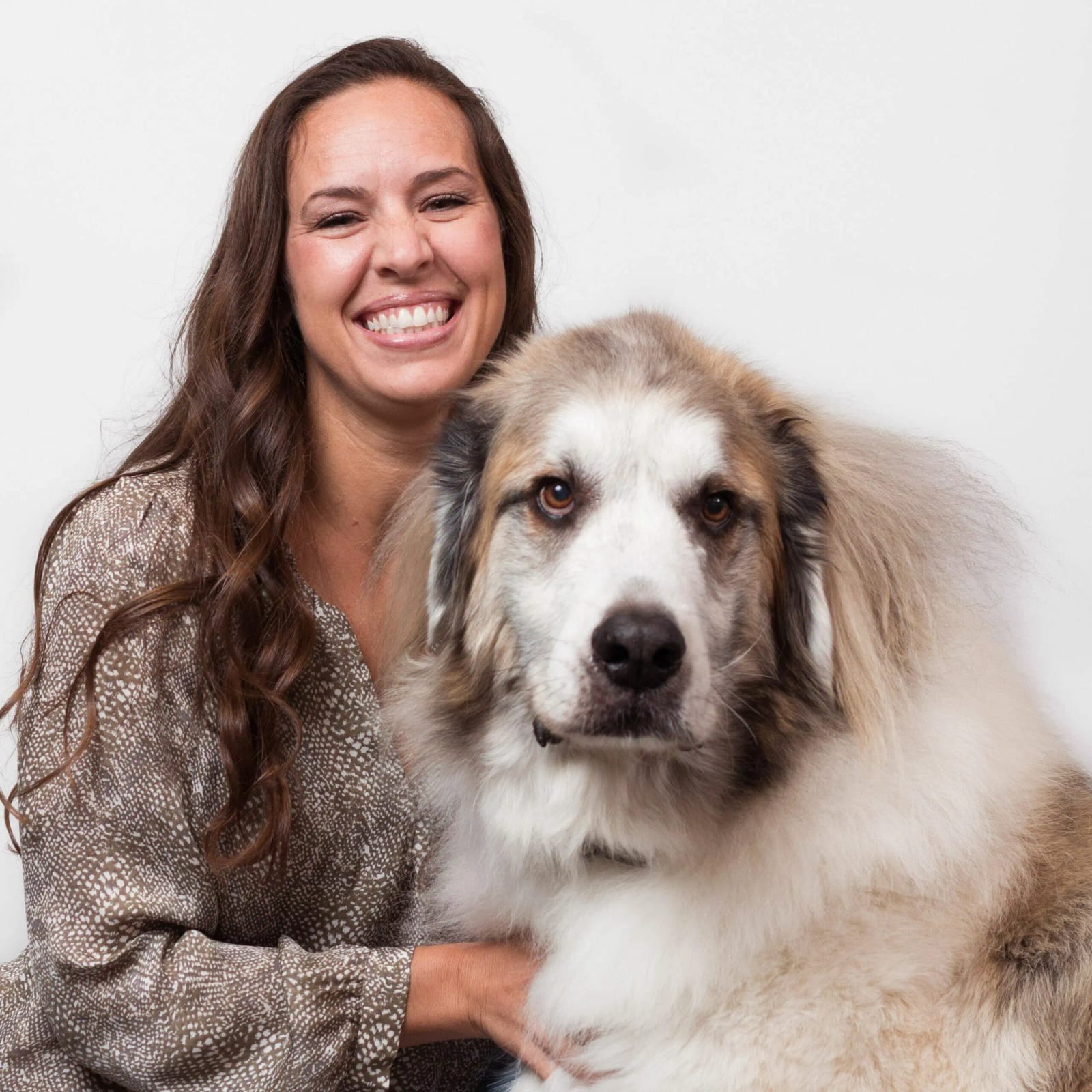 Smiling woman with long wavy hair beside a large fluffy dog against a white background