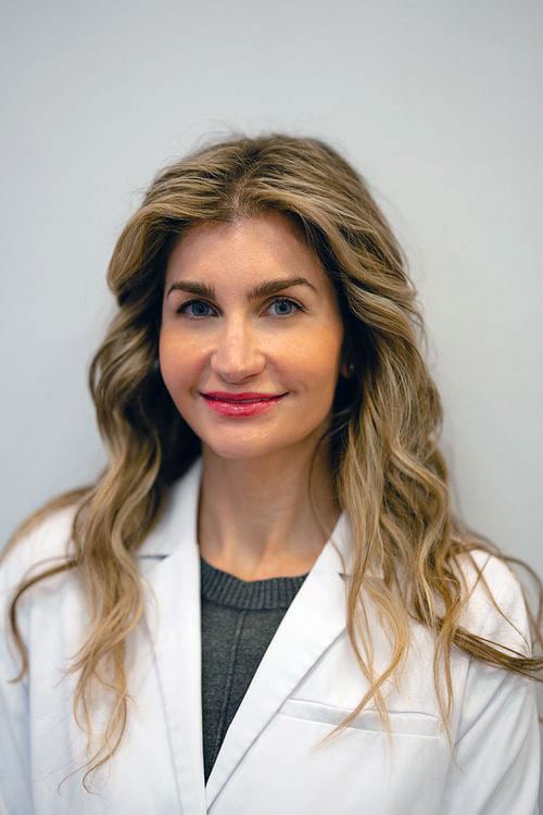 Woman with long wavy light brown hair in a white coat against a plain light wall