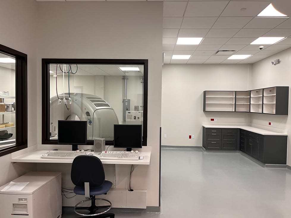 Control area with two monitors beside a window into a room with a large scanner