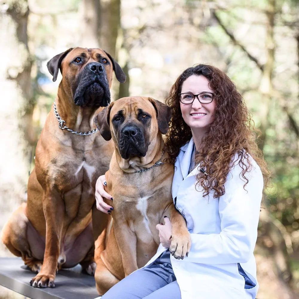 Woman with glasses and curly hair in a white coat sitting with two large tan dogs outdoors