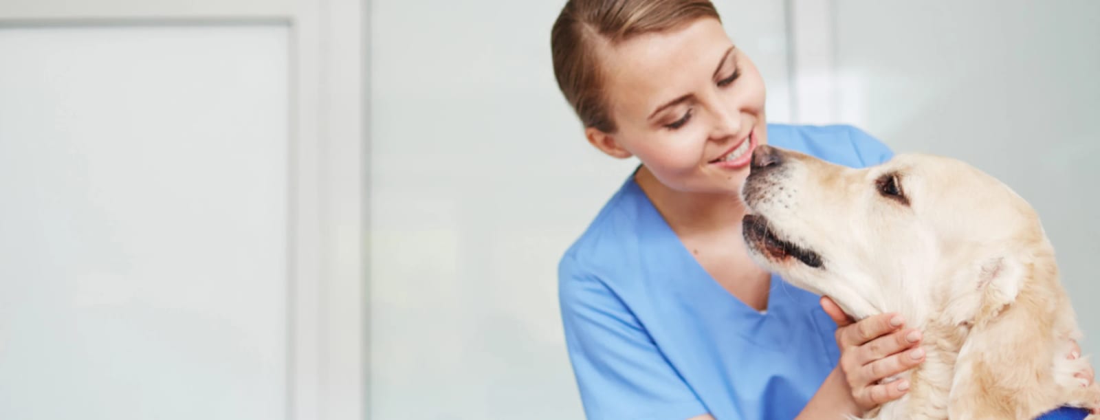 Smiling woman in blue scrubs leaning in as a pale golden dog touches her face with its nose