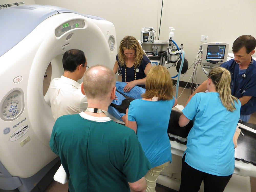 Five people in scrubs gathered around a large white scanner with a patient on the table