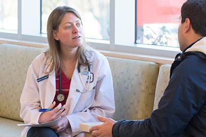 Woman in a white coat with a stethoscope talking with a man on a beige couch