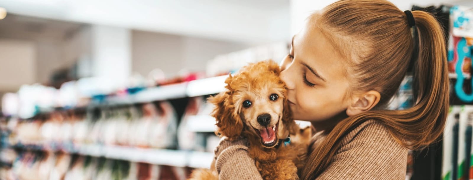 Girl kissing a fluffy apricot puppy in a shop aisle with shelves of products