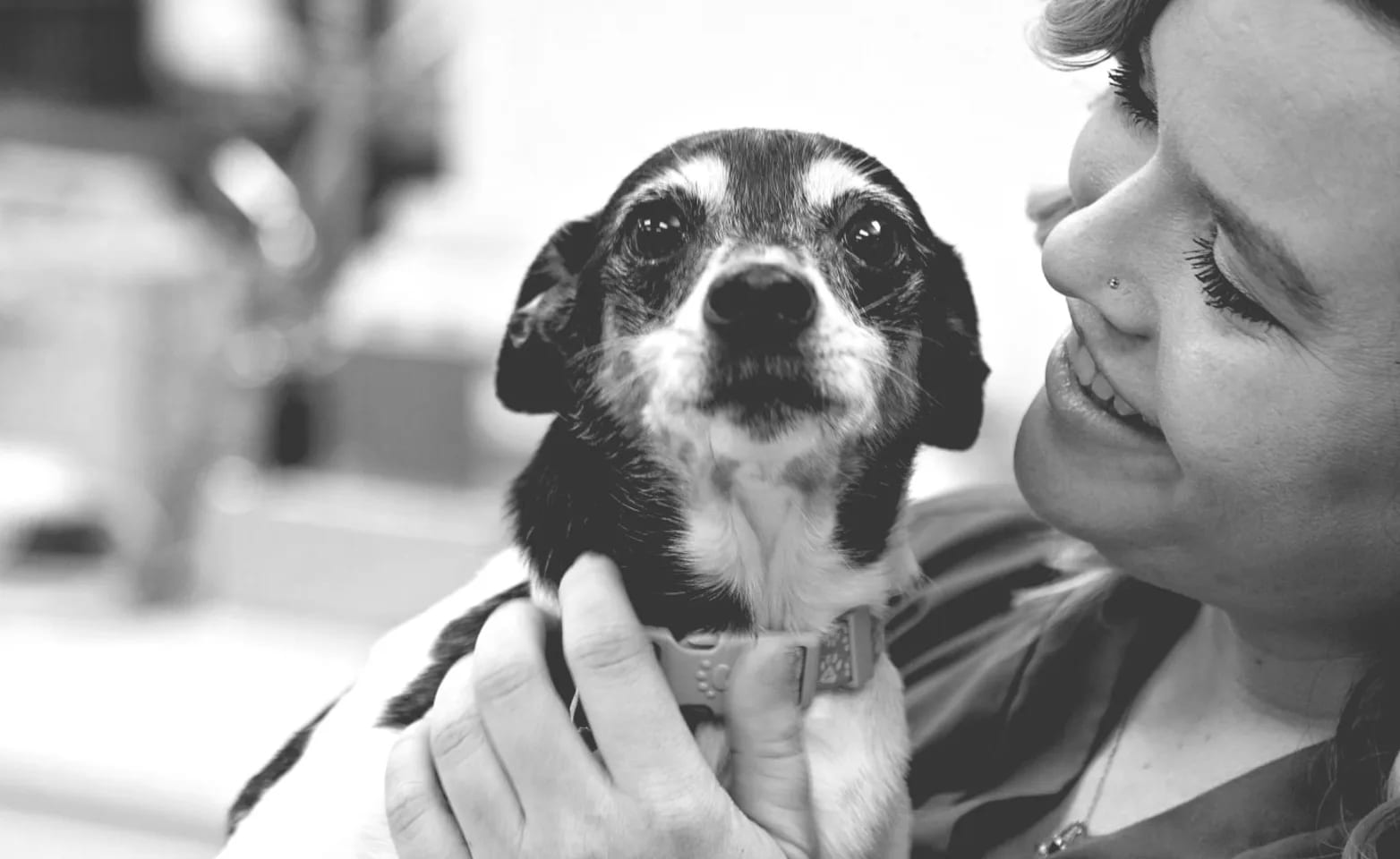 Black and white photo of a smiling woman holding a small dog close to her face