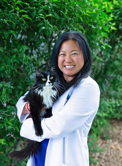 Smiling person in a white coat holding a black and white cat in front of green foliage