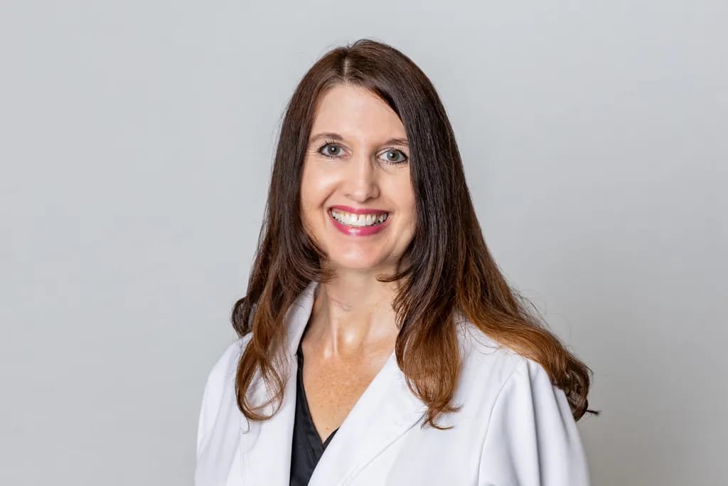 Smiling woman with long brown hair wearing a white coat against a grey background