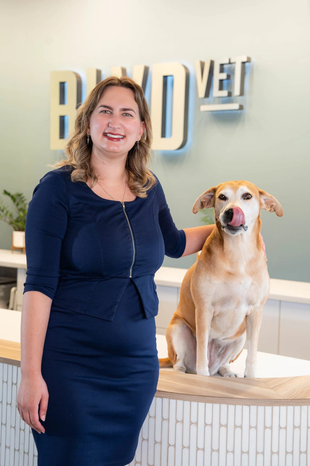 Smiling woman in a navy dress standing beside a tan dog licking its nose on a reception counter