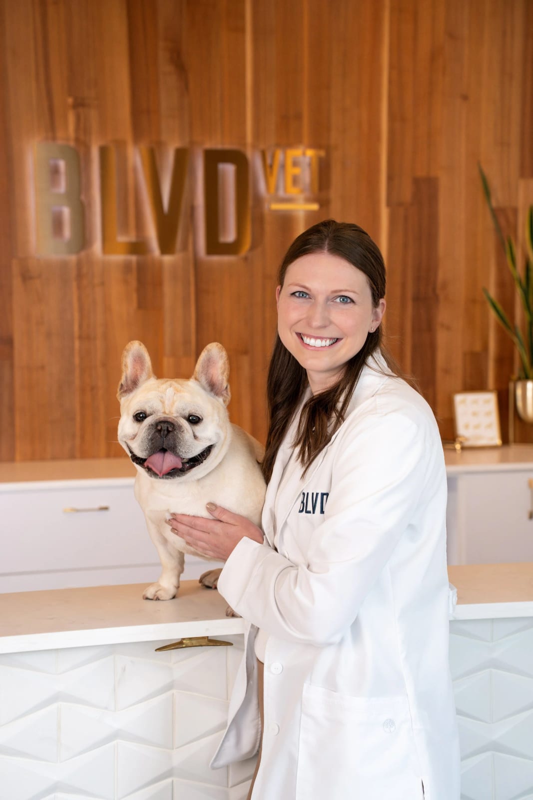 Smiling woman in a white coat with a small cream dog on a counter in front of a wood wall
