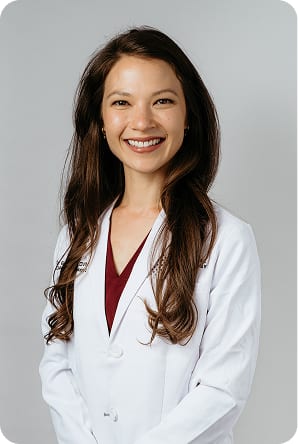Smiling woman with long wavy brown hair in a white coat over a maroon top