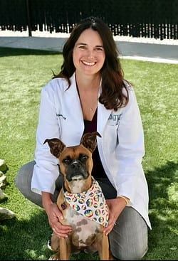 Smiling person in a white coat kneeling on grass with a brown dog in a patterned bandana