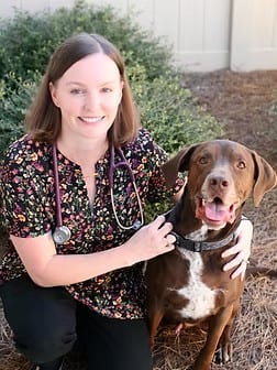 Smiling woman with a stethoscope around her neck crouching next to a brown and white dog