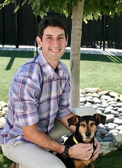 Smiling man in a plaid shirt crouching beside a small dog under a tree