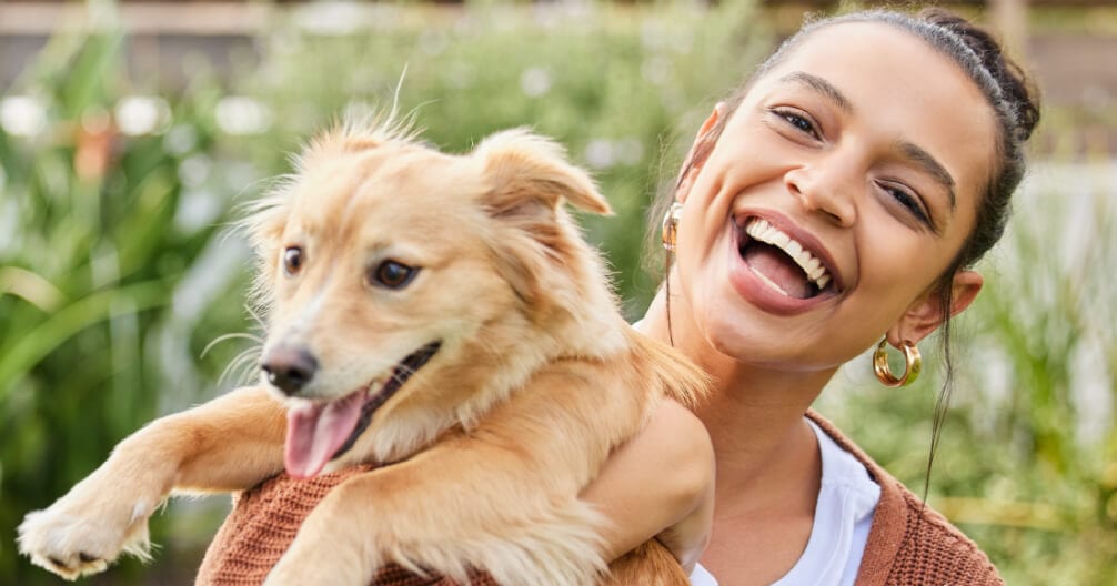 Laughing woman with a hair bun holding a golden-tan dog, with green plants behind