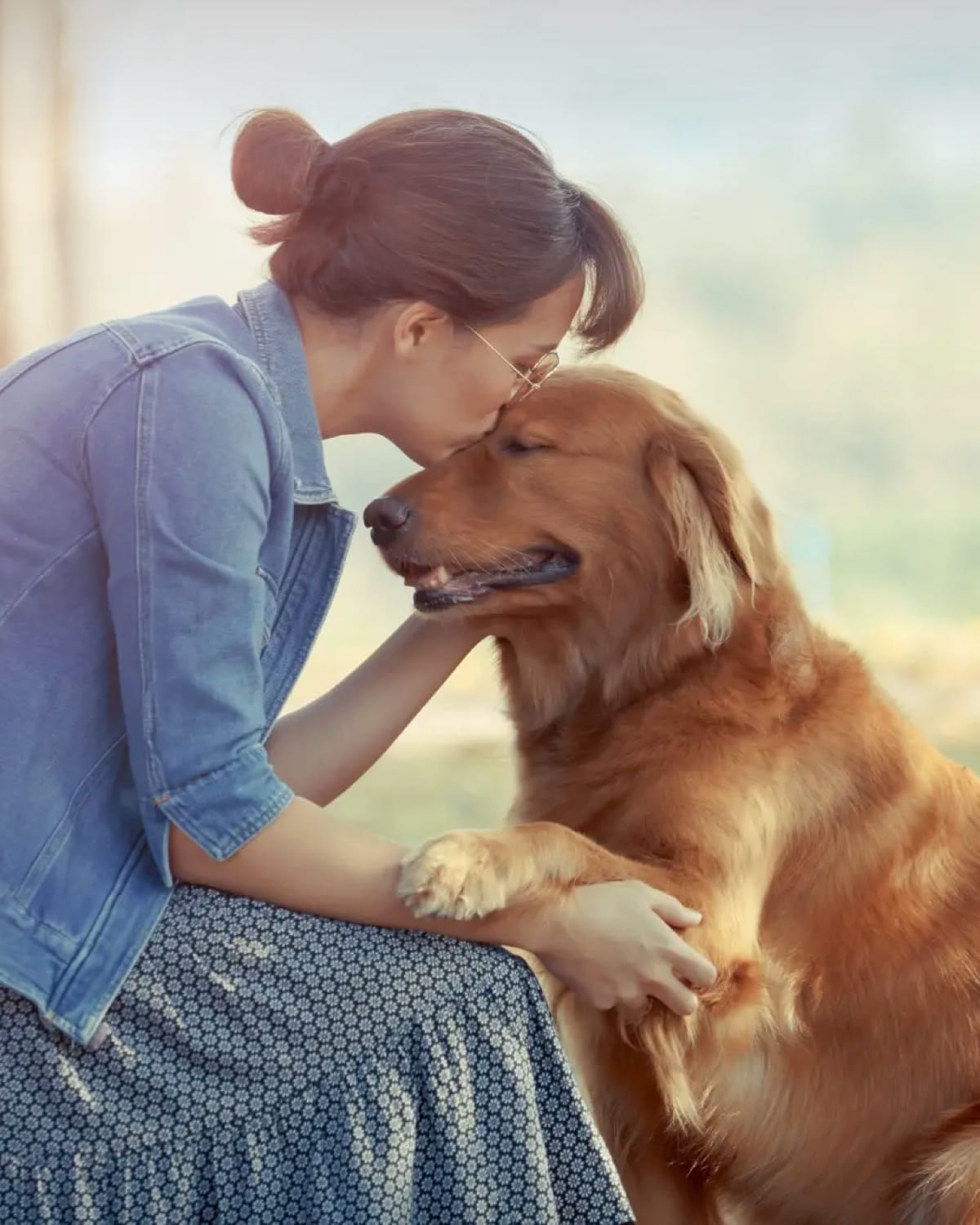 Woman in a denim jacket kissing the forehead of a golden-coloured dog outdoors