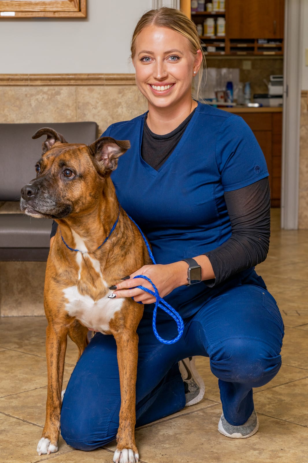 Smiling woman in navy scrubs kneeling beside a brown striped dog on a blue leash