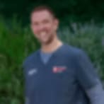 Very small blurry image of a smiling man in a dark blue shirt in front of green foliage