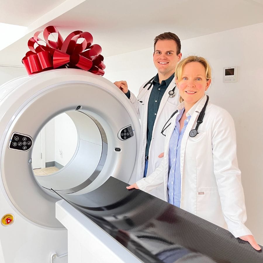 Man and woman in white coats with stethoscopes beside a large white scanner topped with a red bow