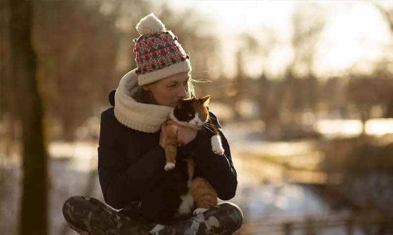Person in a knit hat and dark coat holding an orange, white and black cat to their face outdoors