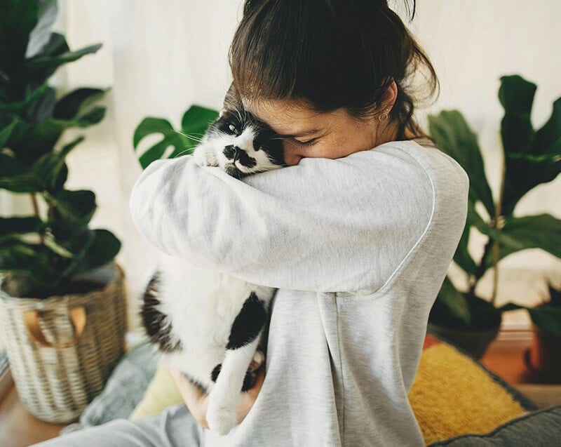 Woman in a grey sweatshirt hugging a black and white cat among green plants