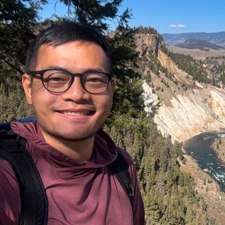 Smiling man with dark glasses and a maroon hooded top taking a selfie on a canyon trail
