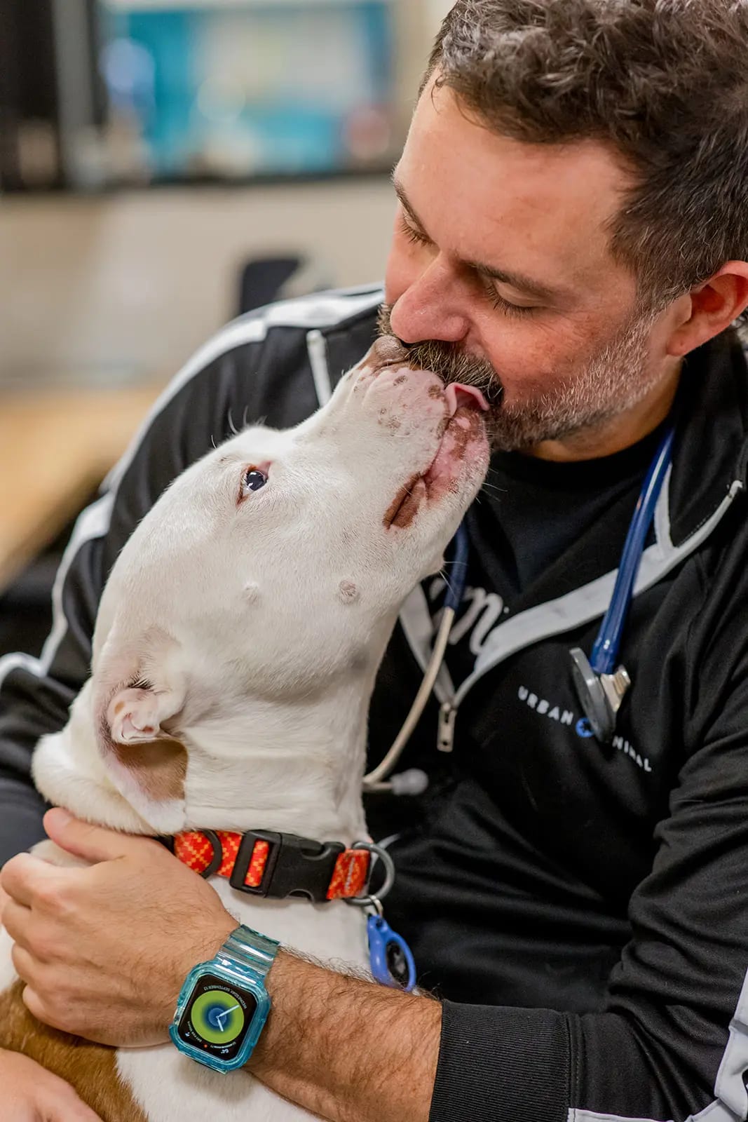 Bearded person in a black jacket with a stethoscope hugging a white dog that licks their face