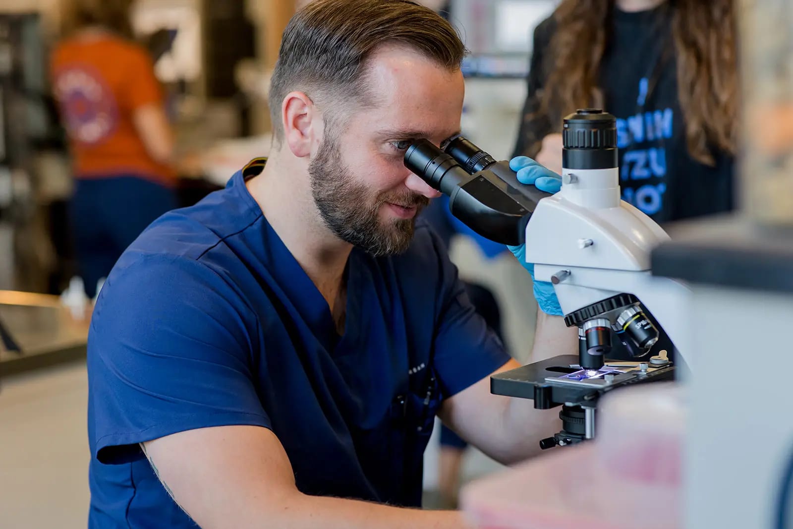 Bearded person in navy scrubs looking into a microscope in a busy room