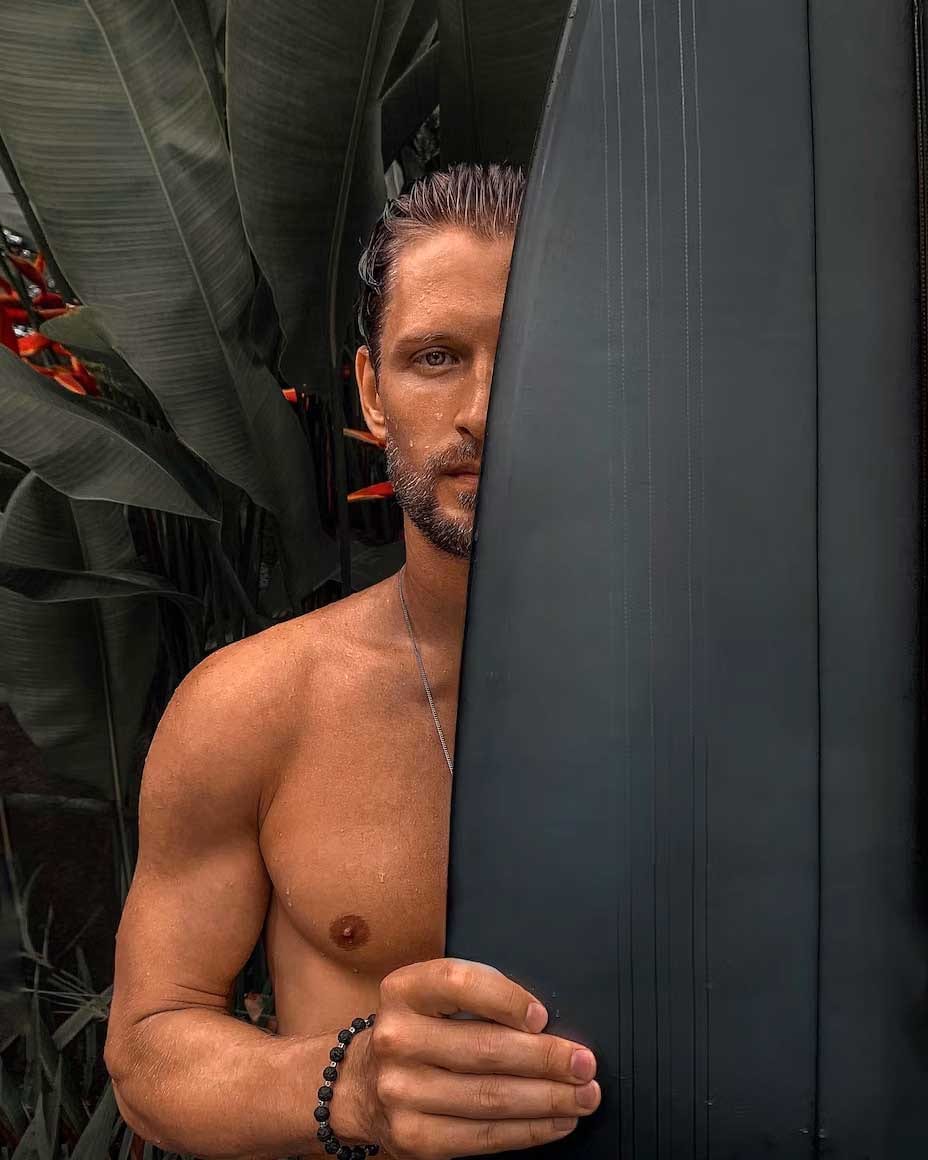 Man with a beard peeking from behind a black surfboard in front of tropical leaves