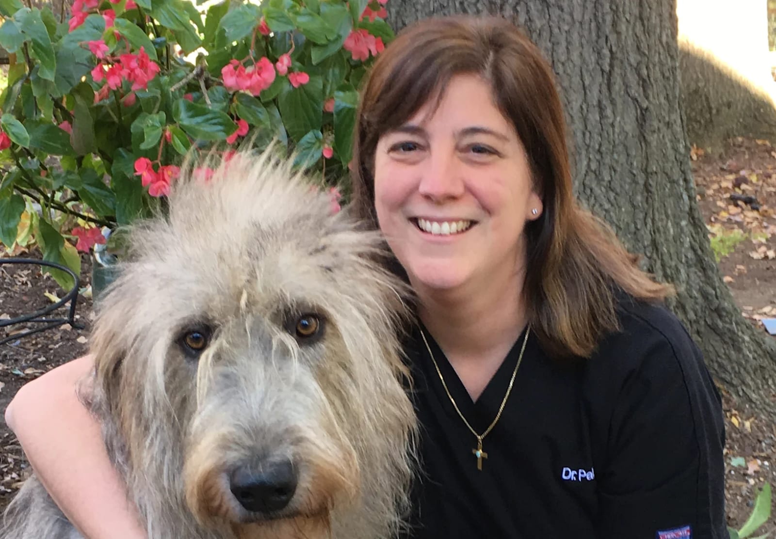 Close view of a smiling person with brown hair and a cross necklace beside a shaggy grey dog