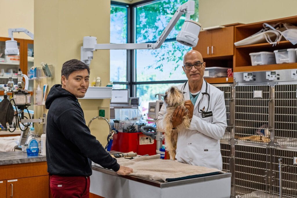 Two men in a clinic treatment room, one in a white coat holding a small fluffy dog next to a steel table