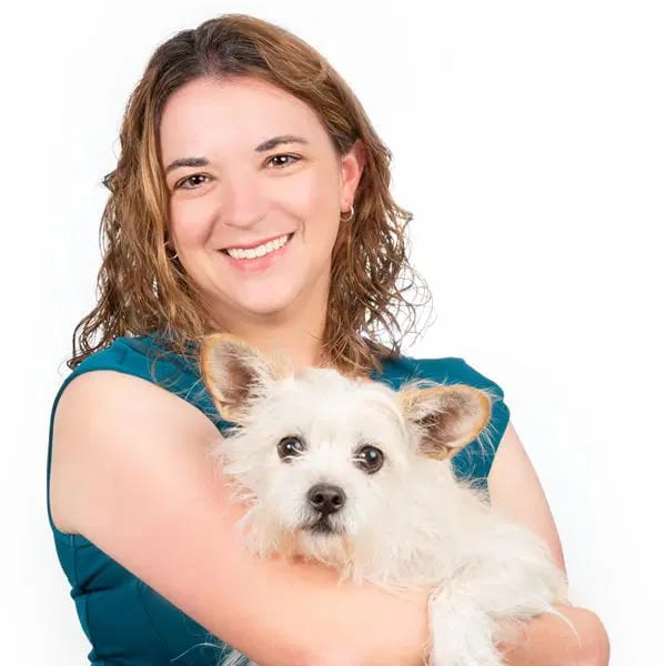 Smiling woman in a teal top holding a small white dog with tan ears against a white background