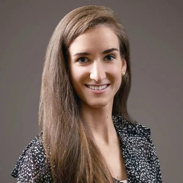 Smiling woman with long brown hair in a black and white patterned blouse against a grey backdrop