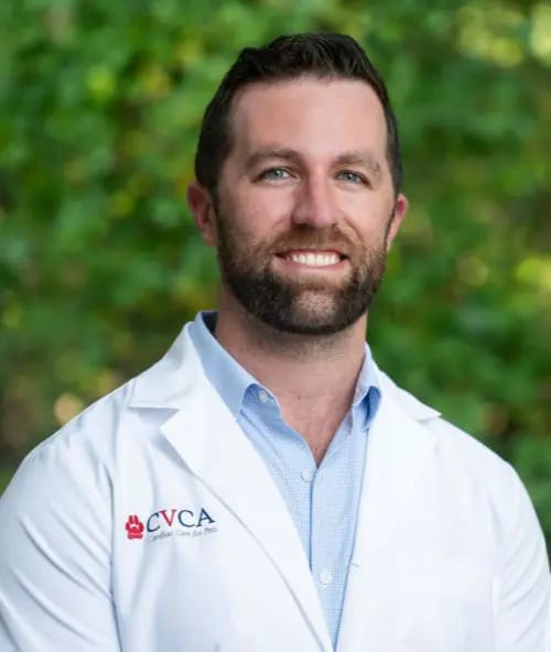 Smiling bearded man in a white coat with a CVCA logo in front of green foliage